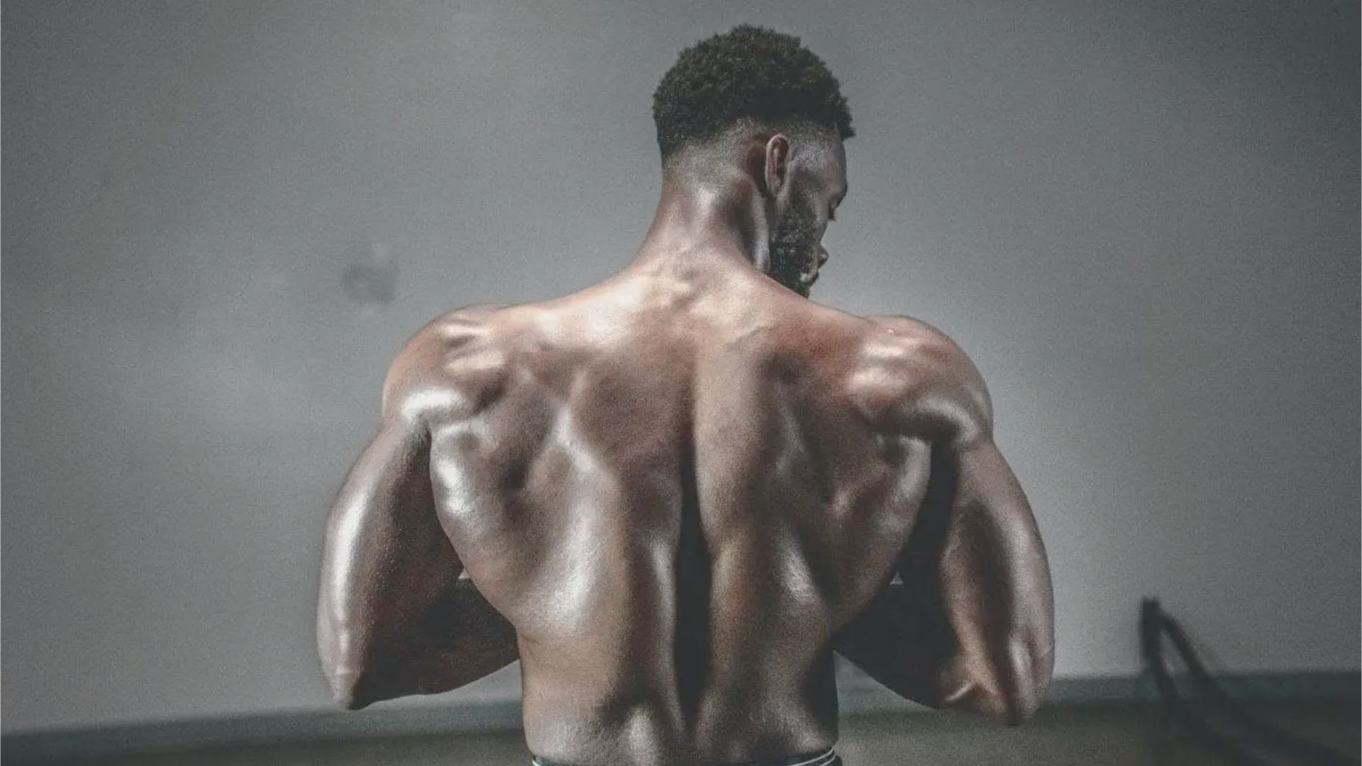 A muscular man with a sculpted back is standing shirtless in a gym, facing away. The dim lighting highlights his defined muscles, conveying strength.