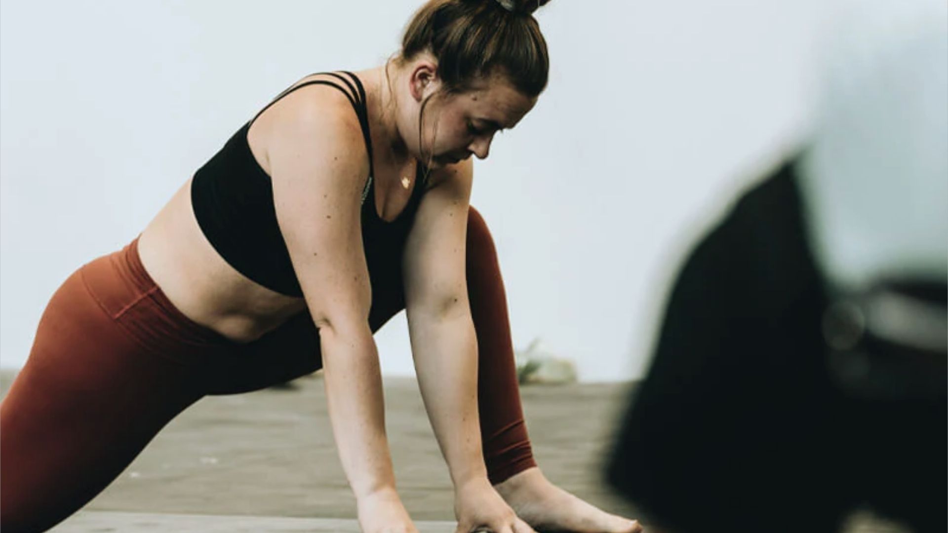 A woman in a black sports bra and red leggings stretches deeply on a yoga mat, focusing intently. The background is softly blurred, conveying calm.