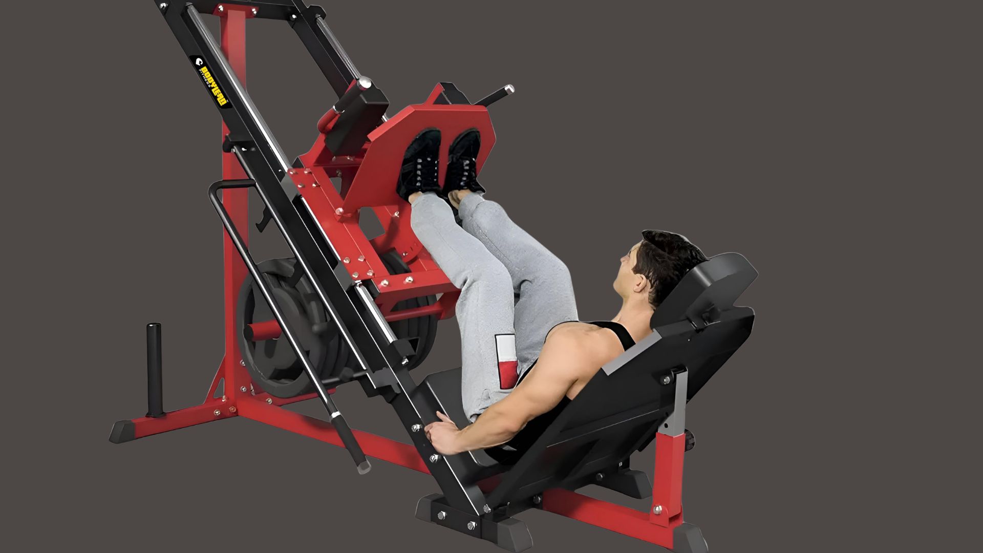 A person uses a red leg press machine in a gym setting, wearing a black tank top and gray pants. The mood is focused and determined.
