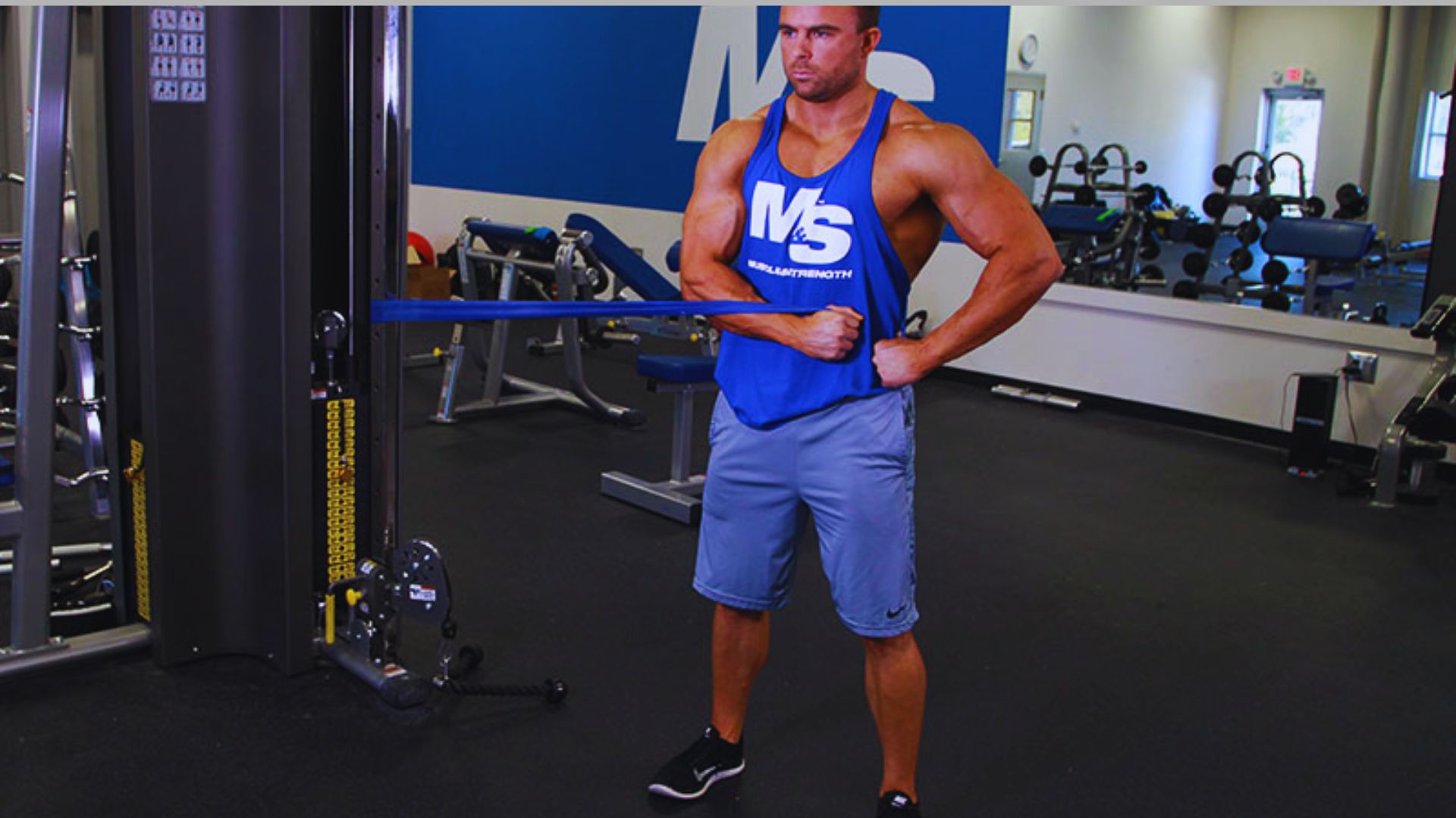 A muscular man in a blue tank top and shorts performs a cable core exercise in a gym. He looks focused, with gym equipment in the background.