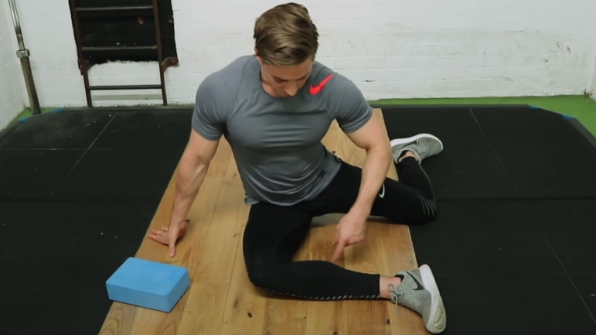 A man in a grey shirt and black leggings performs a seated exercise on a wooden mat, pointing at his bent leg. A blue block is beside him, conveying focus.
