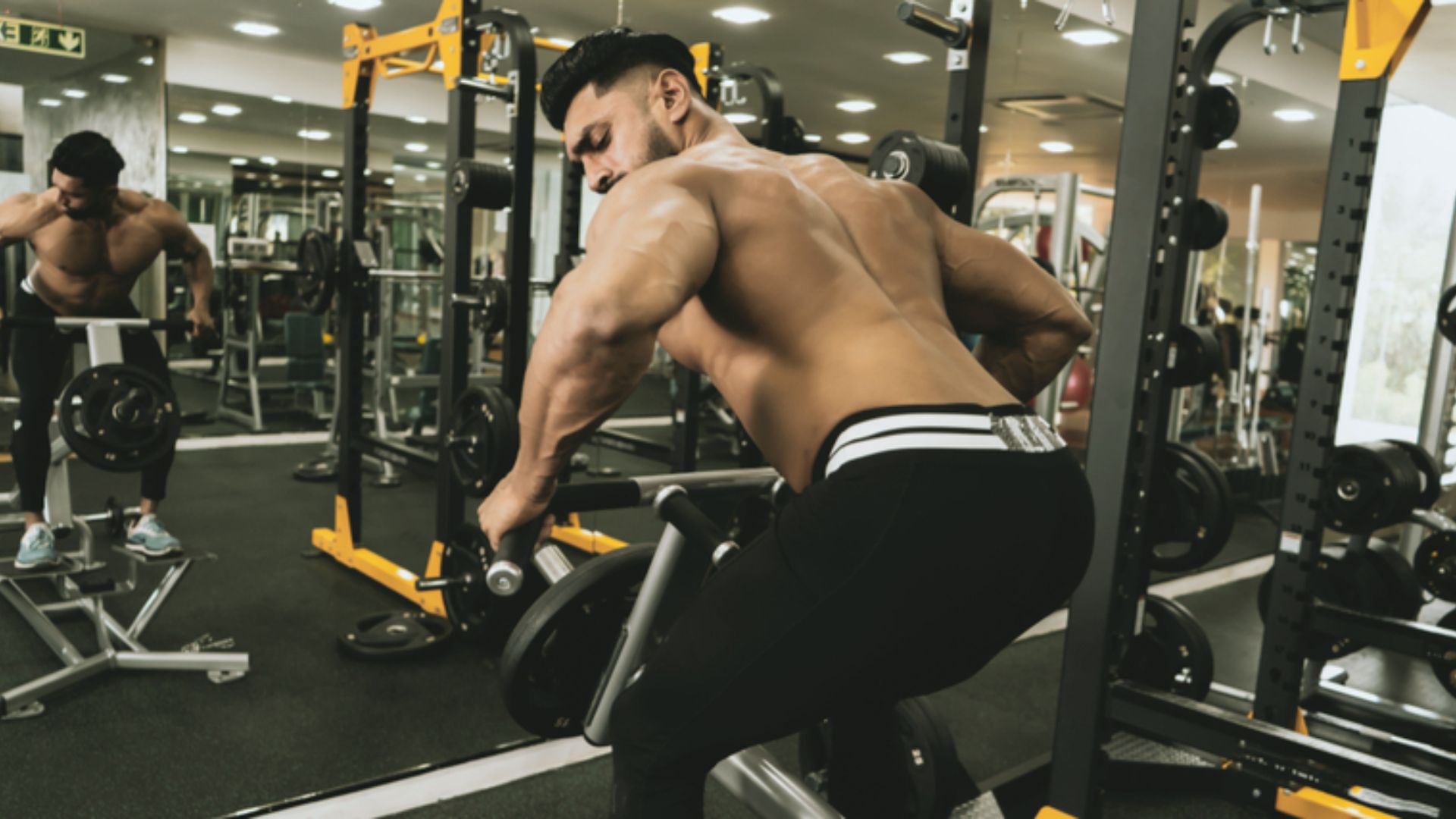 A muscular man performs a bent-over exercise with a barbell in a gym, surrounded by fitness equipment. The scene conveys strength and focus.