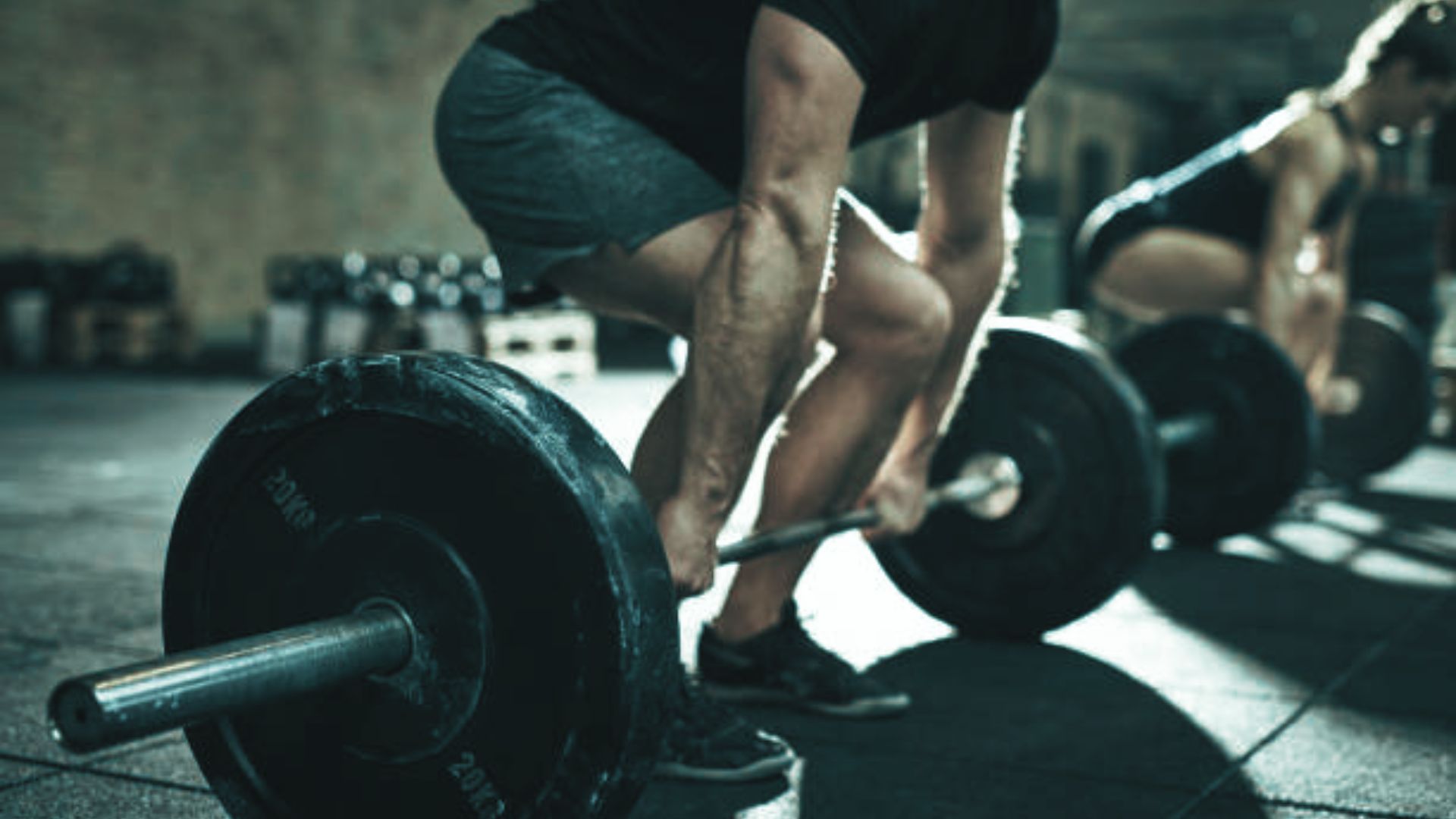 Man and woman lifting heavy barbells in a dimly lit gym. The scene conveys focus and strength, with a gritty, determined atmosphere.
