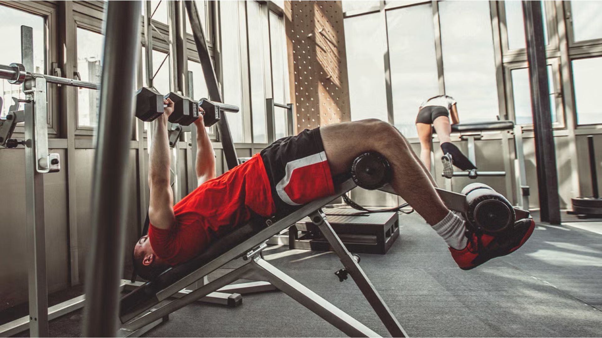 Man in red shirt lifts dumbbells on an incline bench at the gym, while another person exercises in the background.