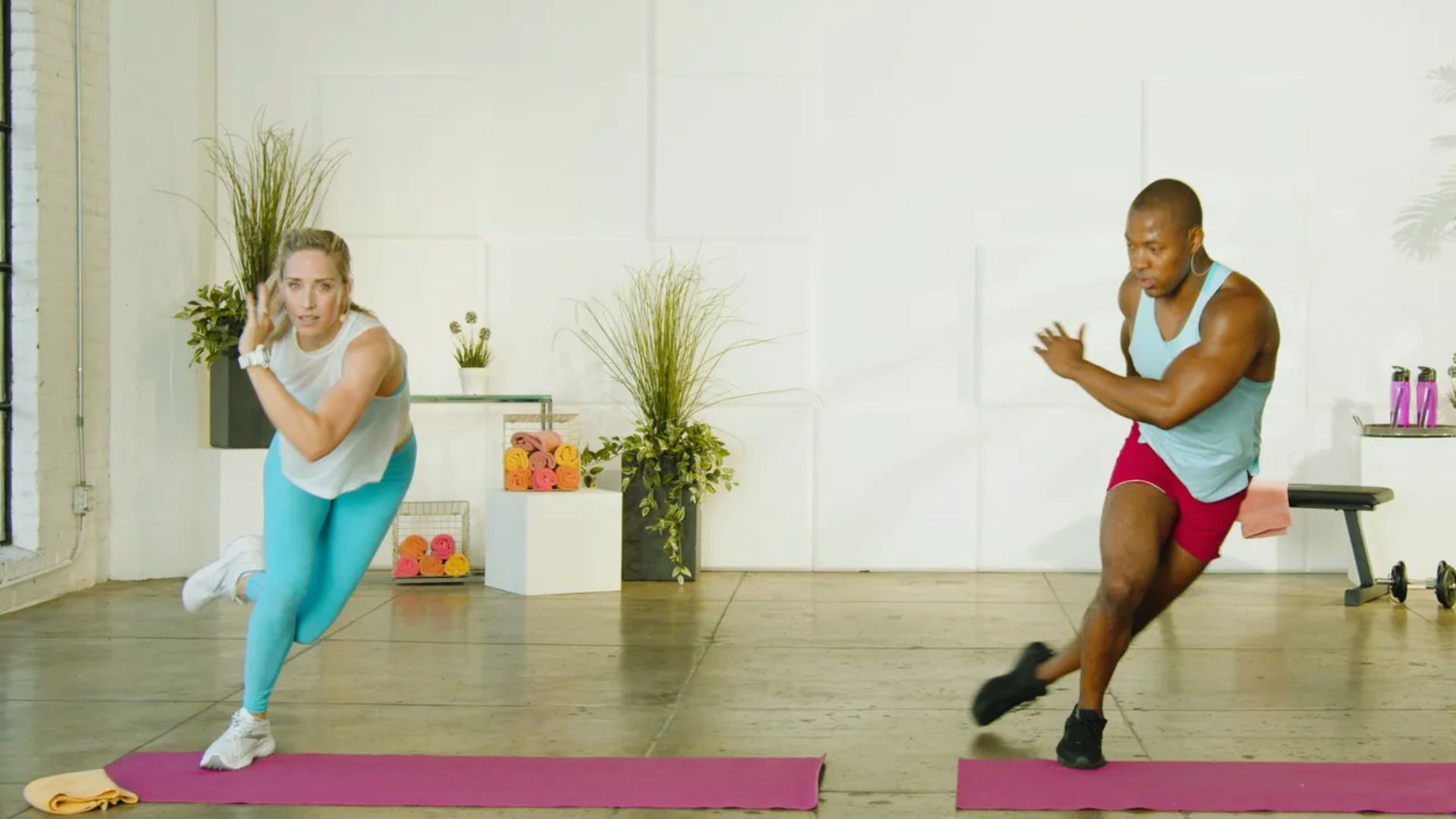 Two people exercise in a bright studio, doing lateral lunges on pink mats. The setting includes plants and colorful boxes, creating an energetic feel.