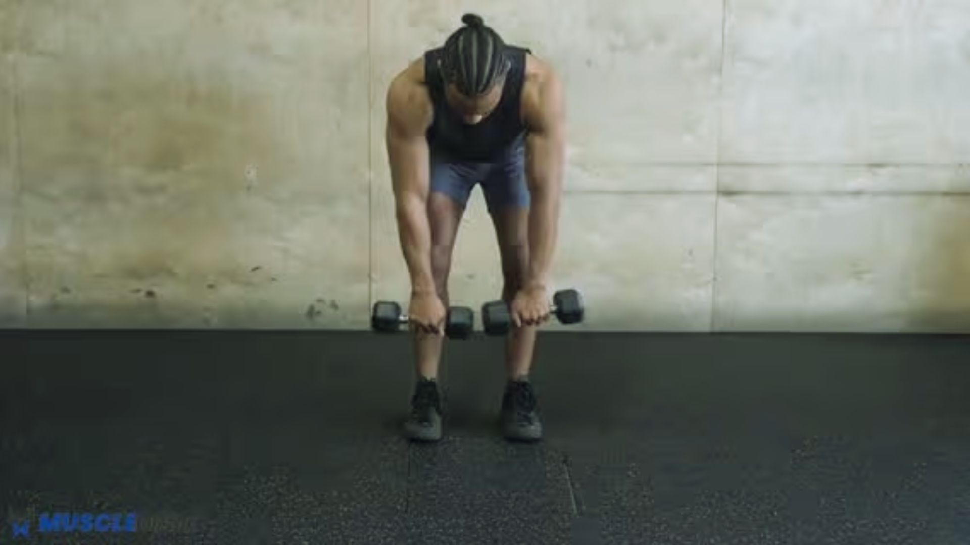 A person in workout attire performs a bent-over dumbbell exercise in a gym. They are focused and determined, with a concrete wall backdrop.