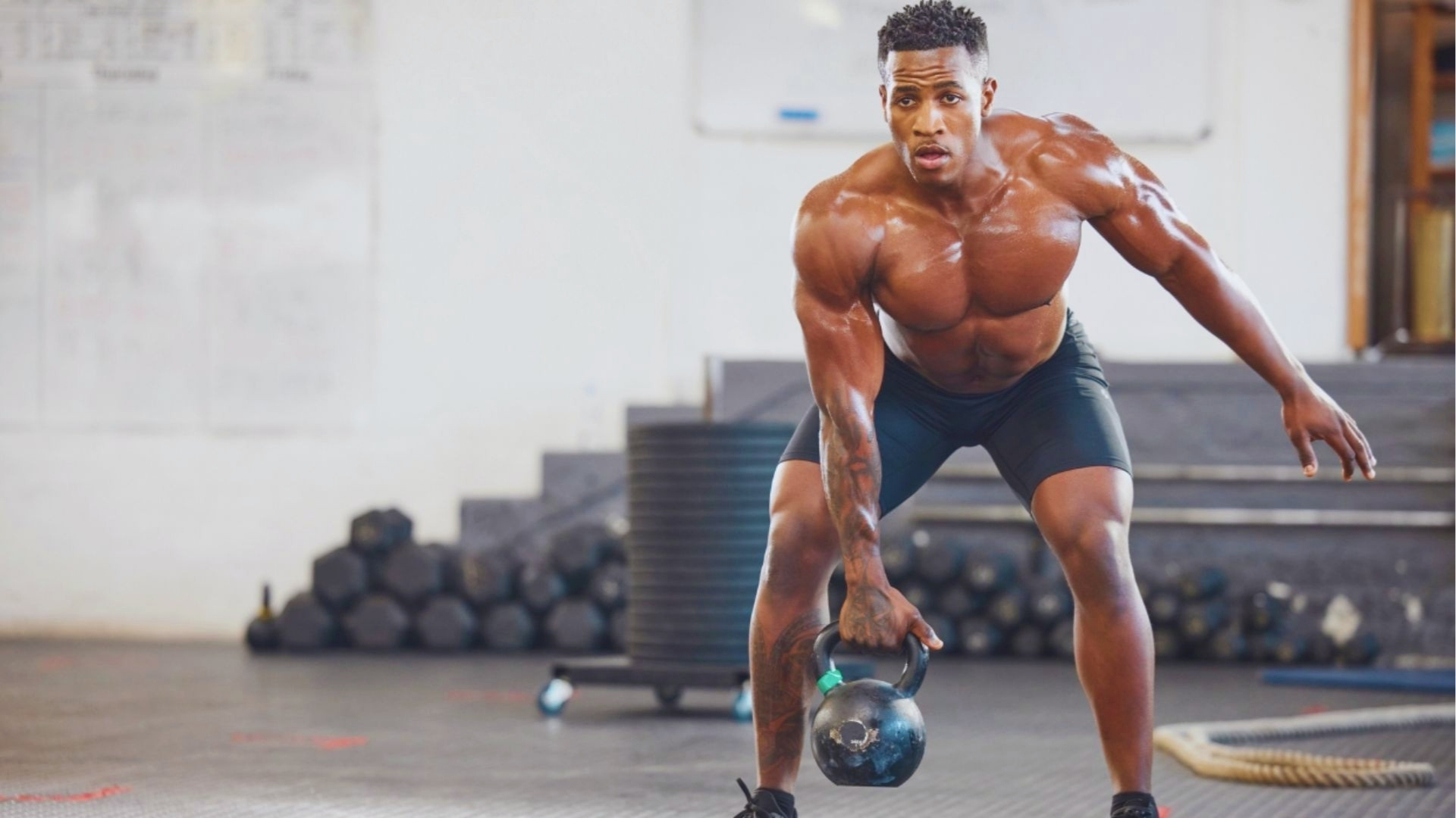 A muscular man exercises in a gym, holding a kettlebell with focus. He is wearing black shorts, and weights are stacked in the background. Energetic mood.