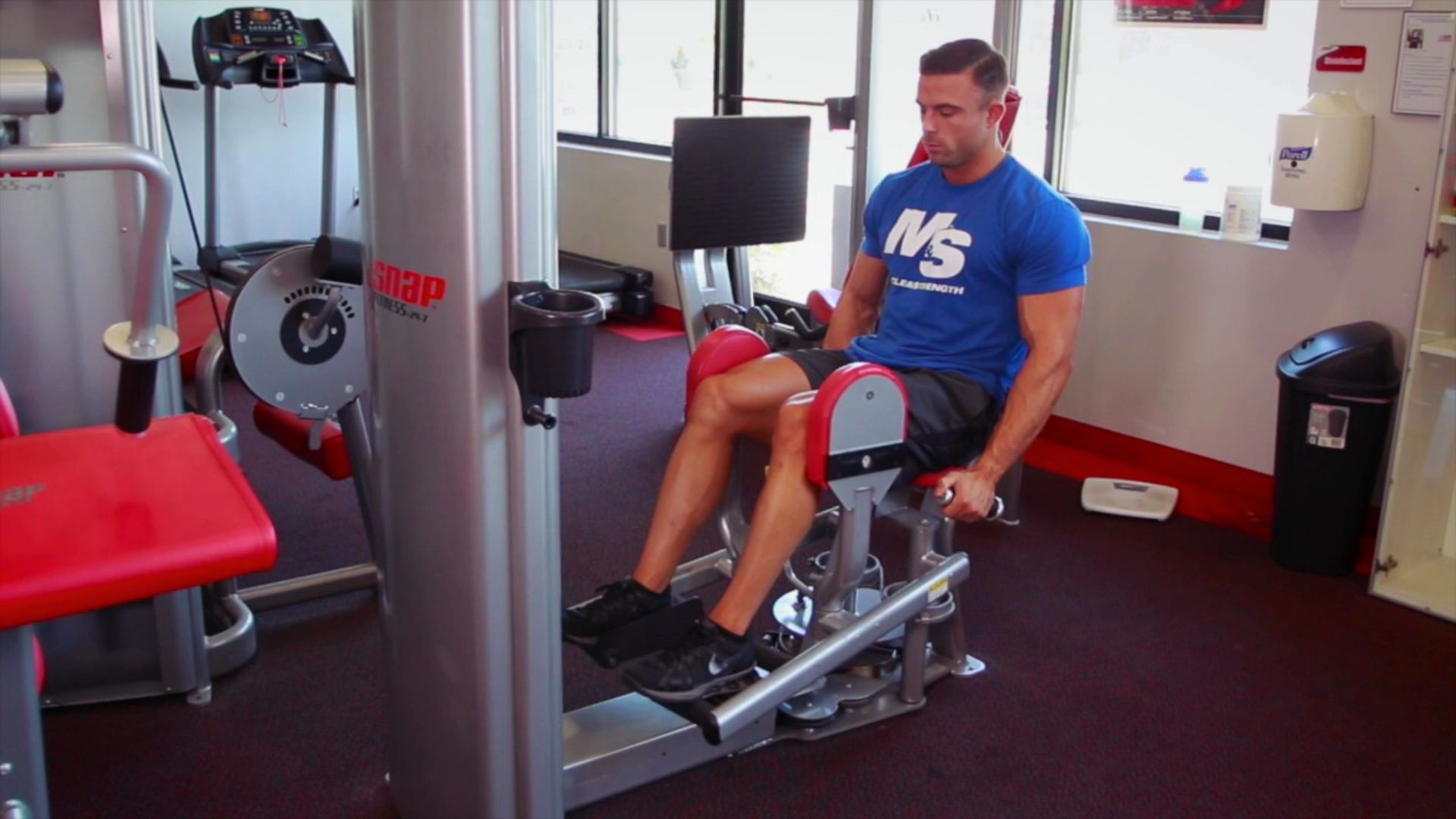 A man in a blue shirt uses a seated leg press machine in a gym. The room is well-lit with large windows, creating a focused yet calm atmosphere.