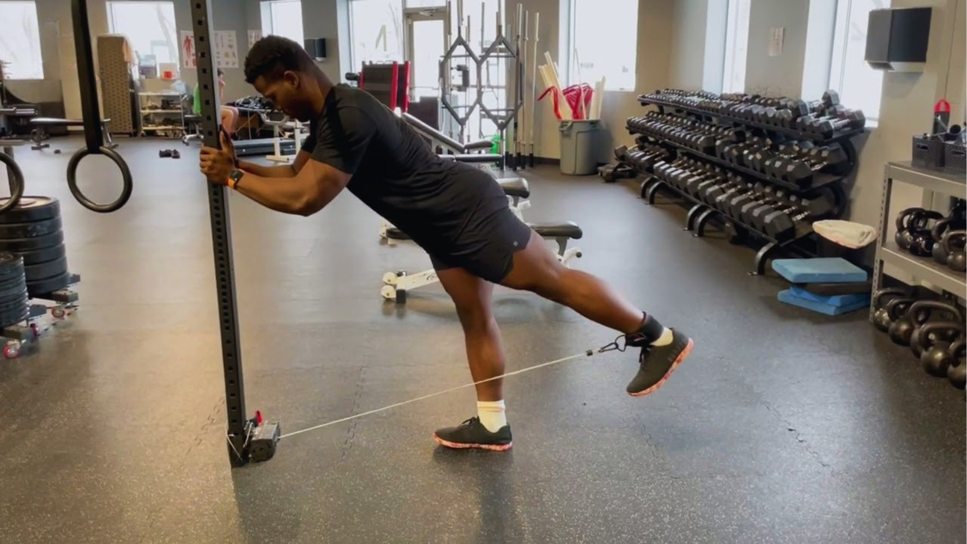 A person exercises in a gym, using a resistance band attached to a rack. They lean forward with one leg extended back. The room is filled with weights.