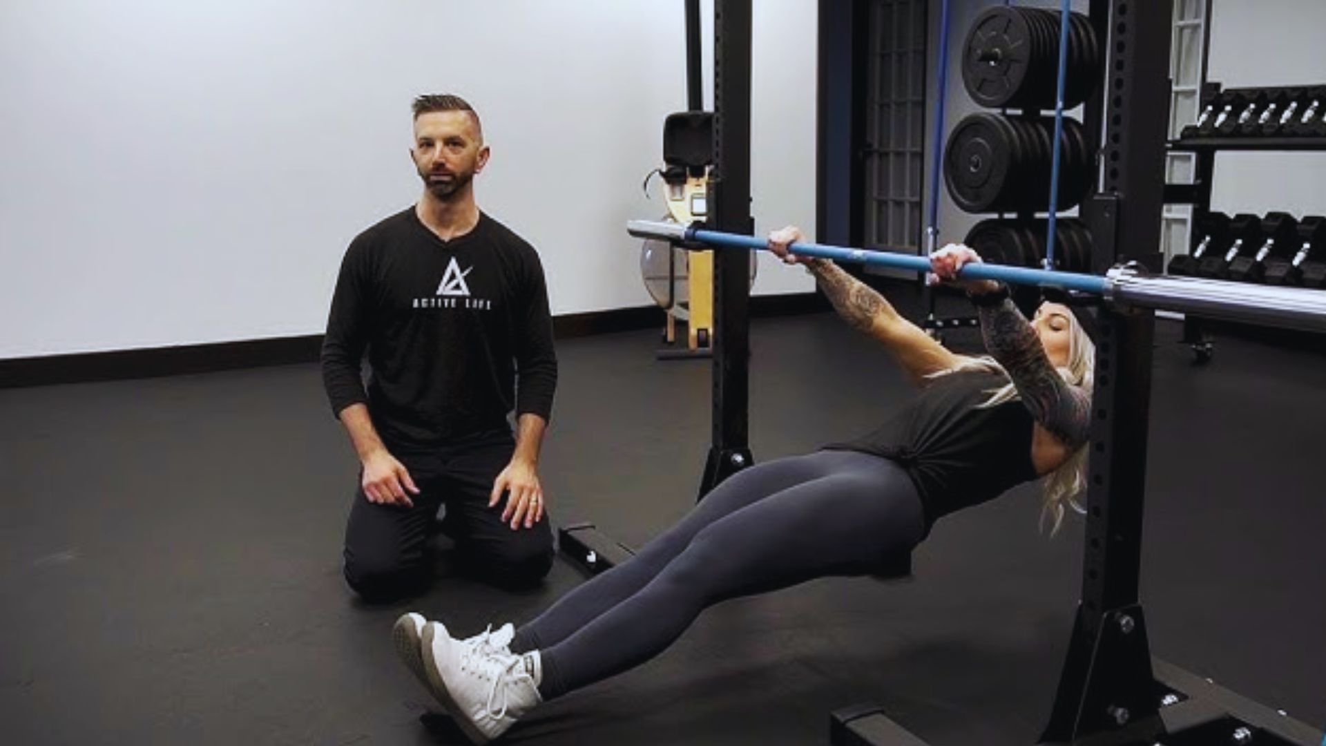 A woman performs an inverted row using a barbell, while a trainer kneels nearby observing. The gym setting is minimalistic and focused.
