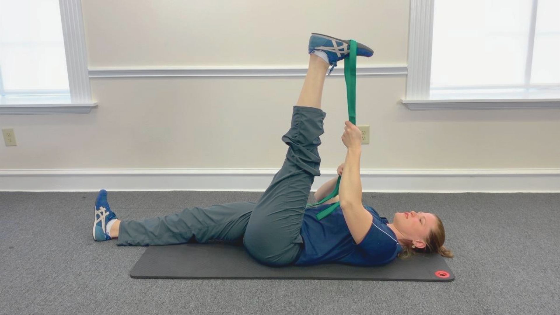 A woman performs a hamstring stretch on a gray yoga mat, pulling her right leg up with a green strap. She's relaxed, wearing blue shoes and clothes.