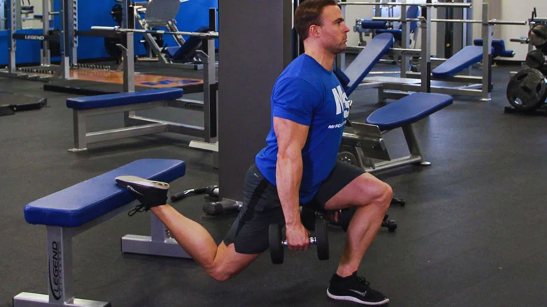 A man in a gym performs a Bulgarian split squat with dumbbells. He's focused, wearing a blue T-shirt and black shorts, with one leg on a bench behind him.
