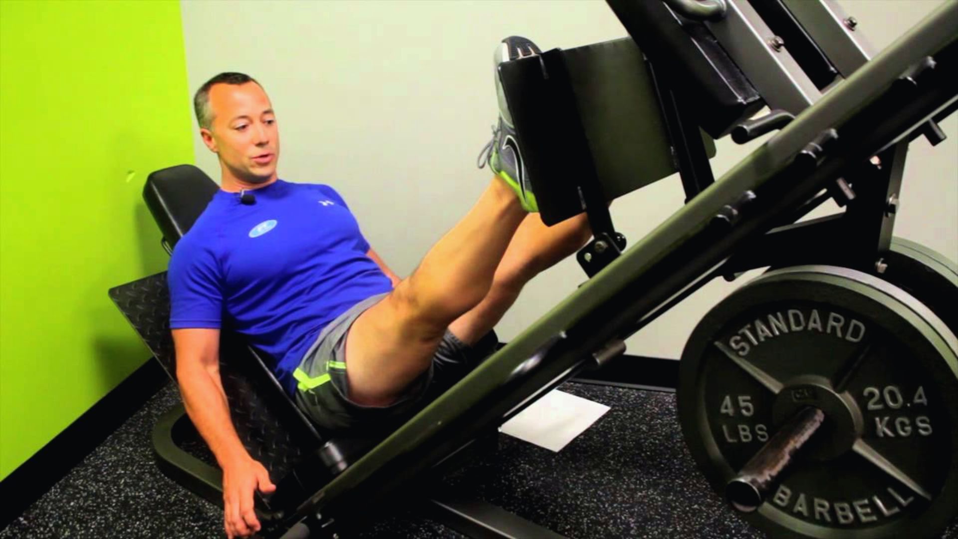 Man in blue shirt performs a leg press in a gym. He is focused, pushing weights with his feet. The room has a bright green wall, creating a motivational atmosphere.