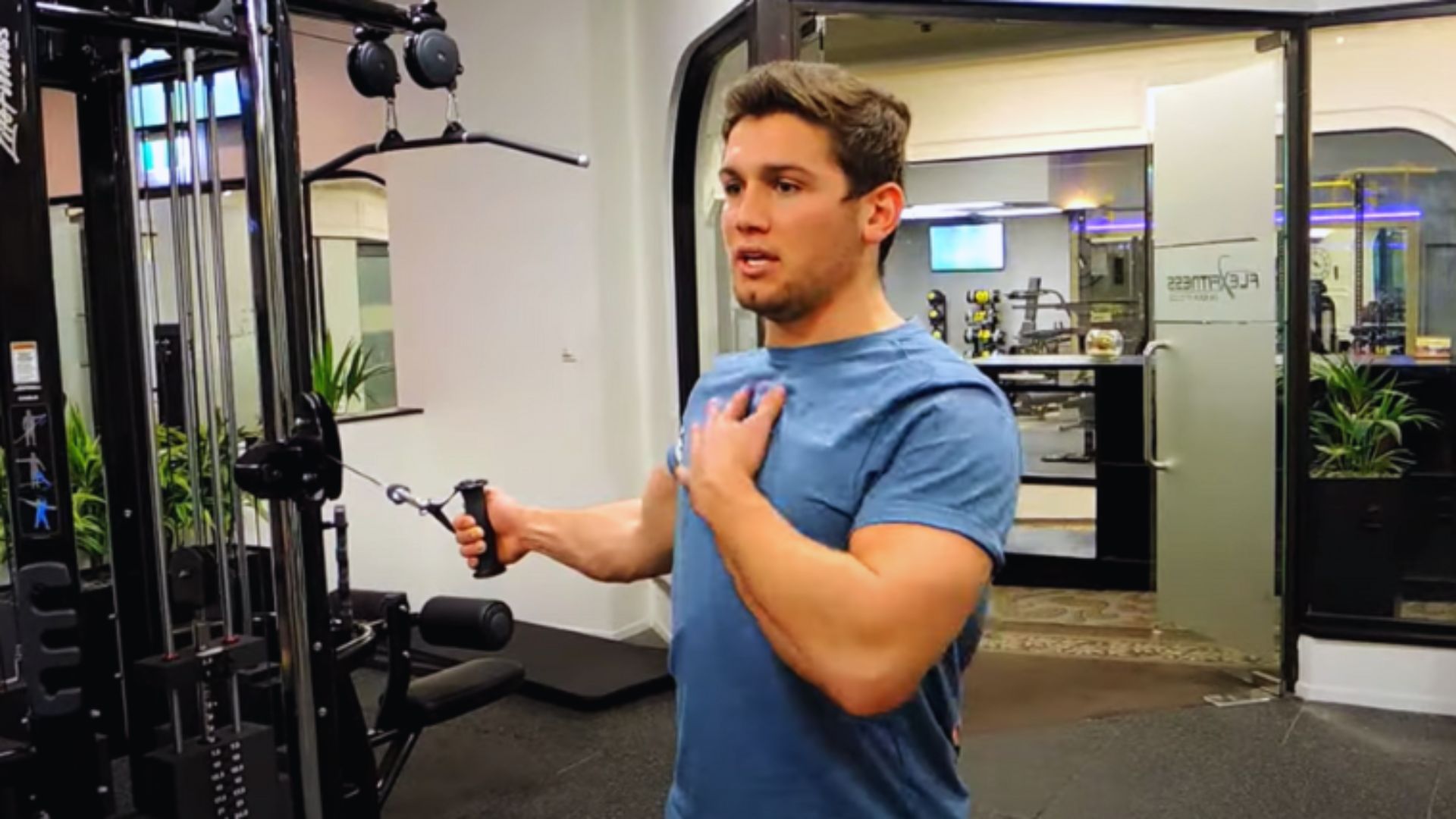 A man in a blue t-shirt uses a cable machine at the gym. He pulls a handle with one arm while touching his chest with the other. The gym is modern and well-lit.