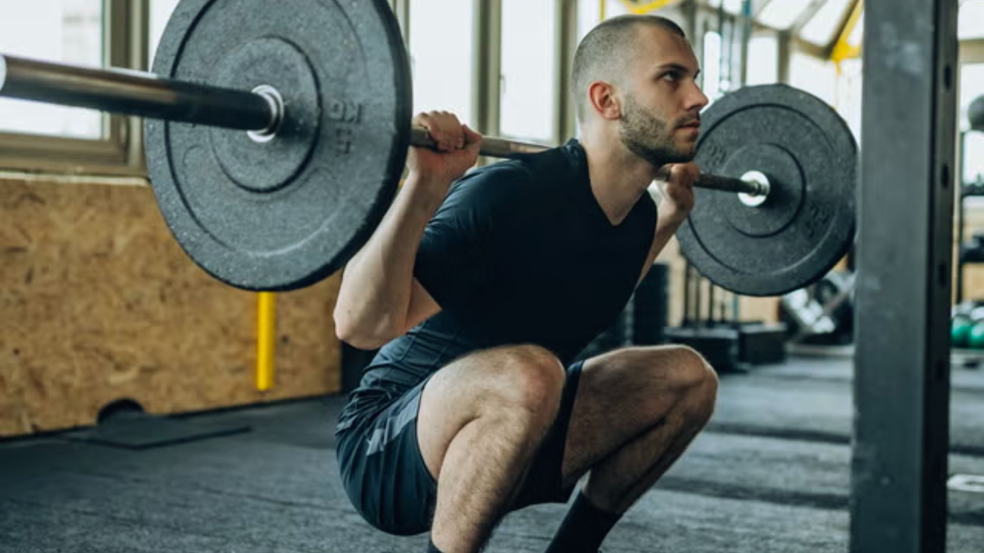 A man in athletic wear performs a barbell squat in a gym, demonstrating focus and strength. Sunlight filters through windows, creating a determined atmosphere.