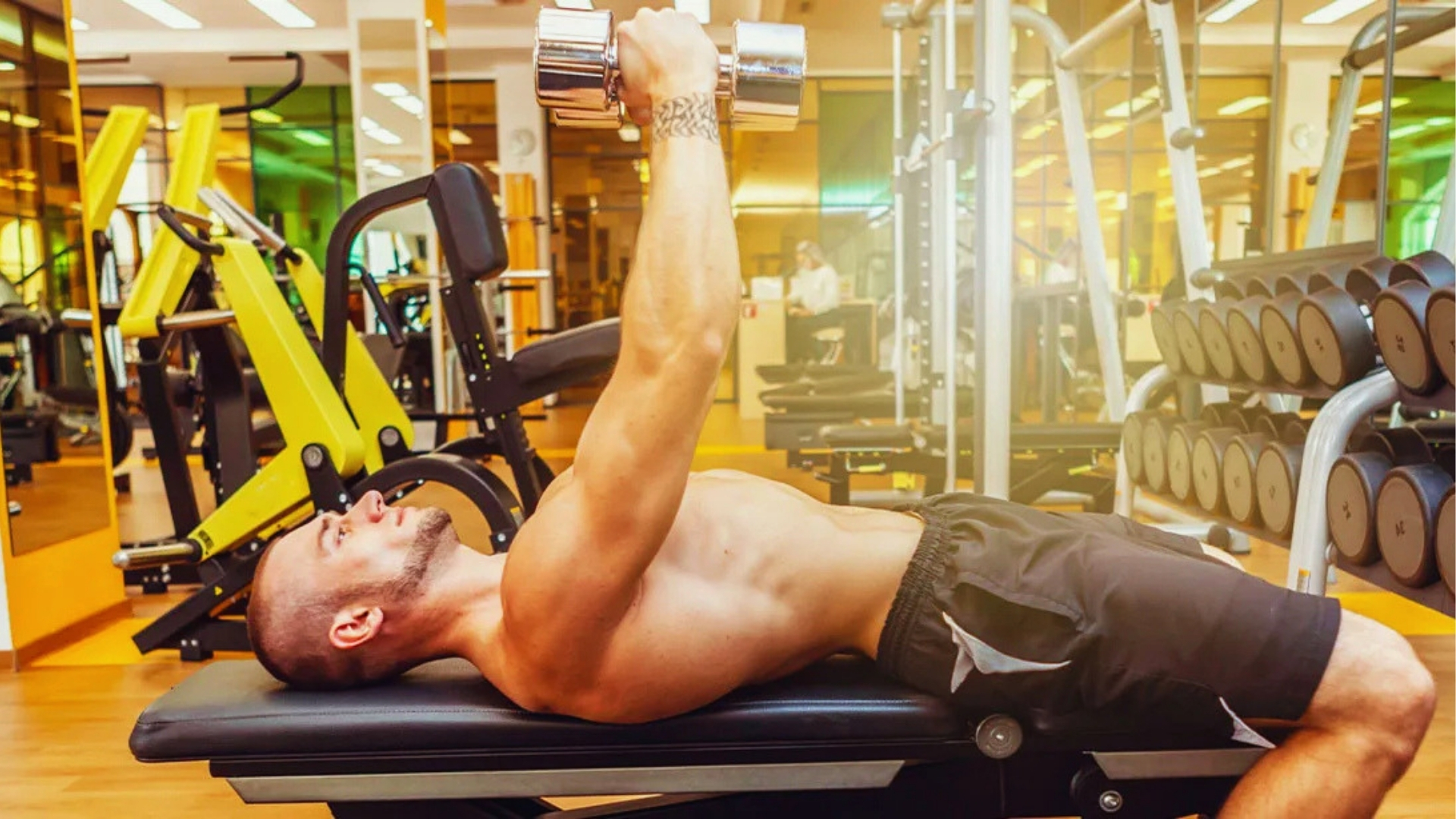 A man at the gym is lying on a bench, lifting a dumbbell with one hand. The room is filled with fitness equipment, creating an energetic ambiance.