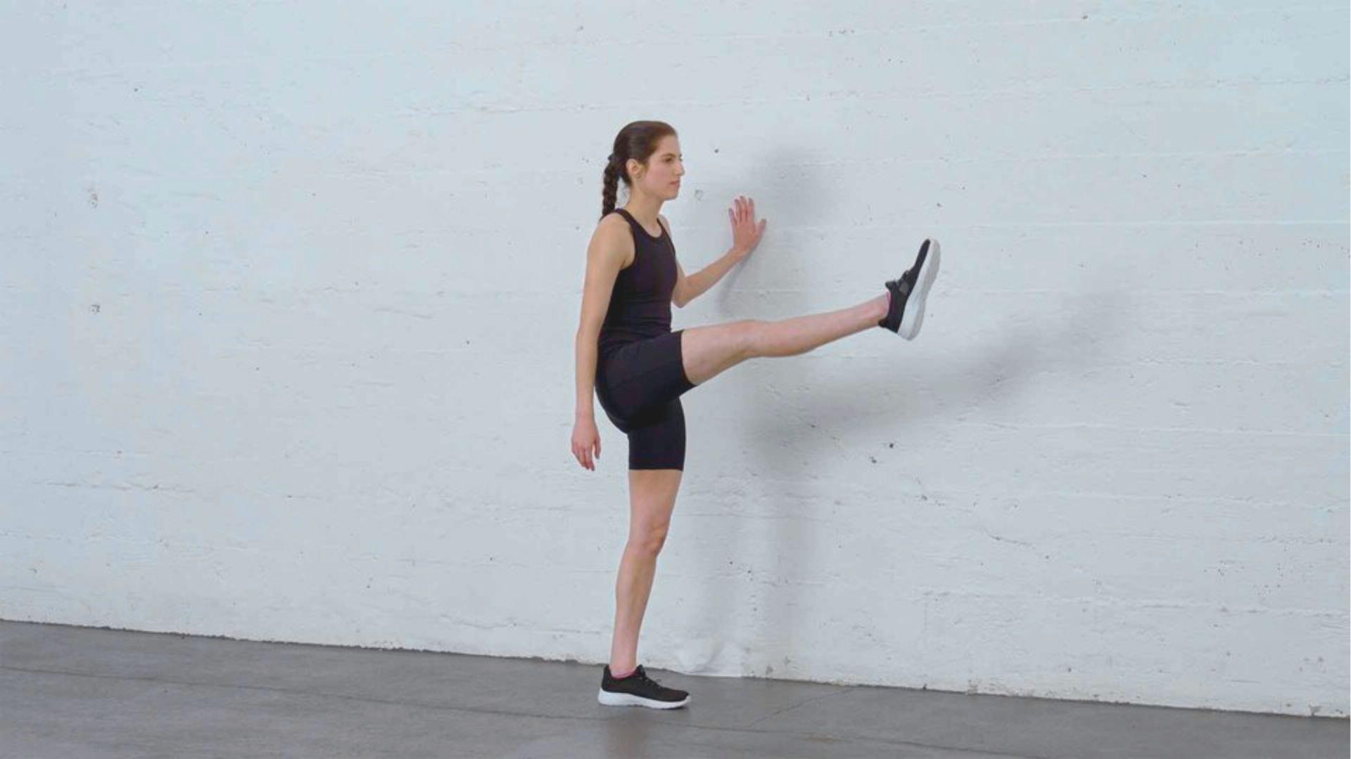 Woman in black workout set performs leg swings against a white wall, hand placed for balance.