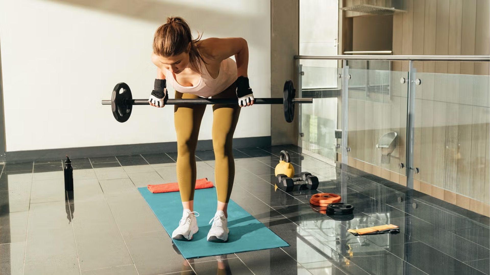 Woman in workout gear does bent over barbell row on yoga mat in bright home gym space