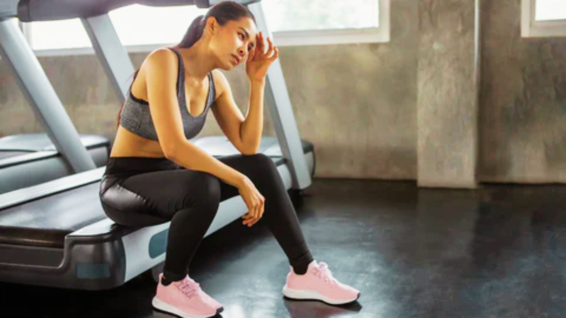 Exhausted woman in grey activewear sits on a treadmill, resting her head on her hand with a tired expression.