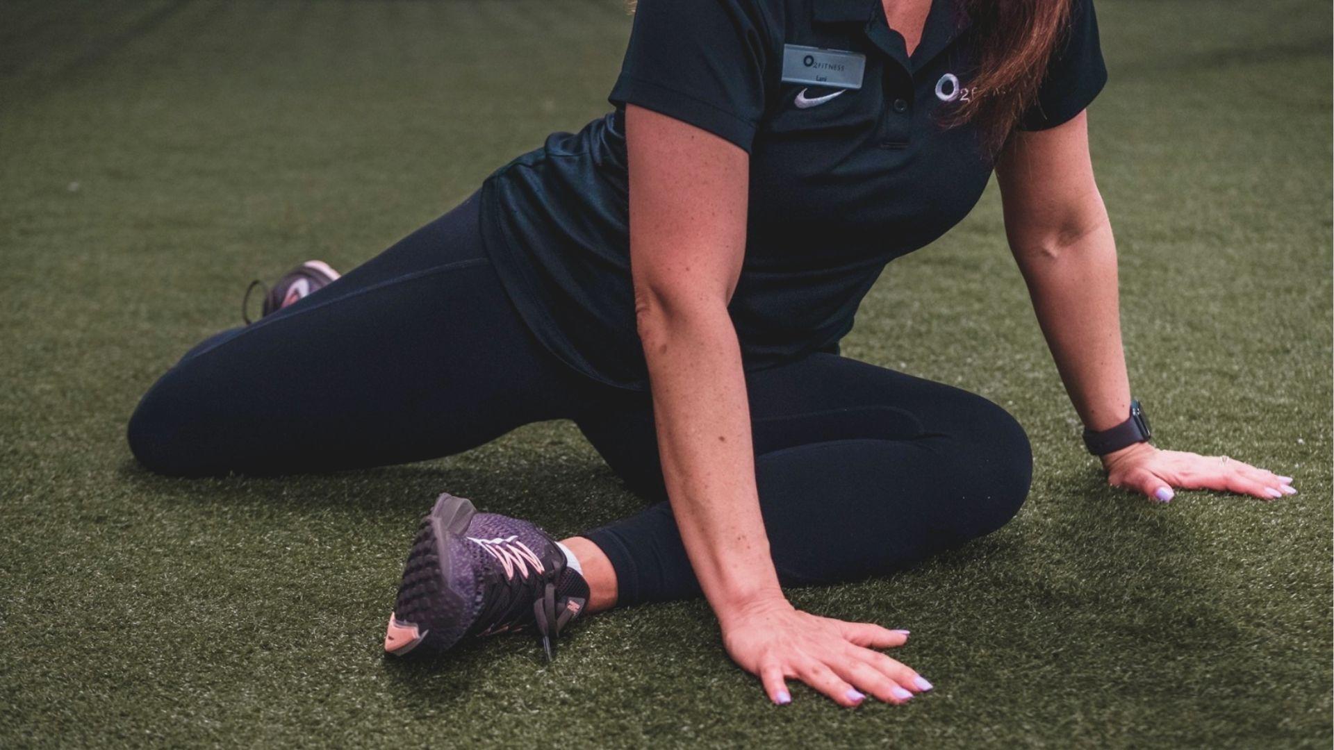 Fitness trainer in black Nike polo demonstrating a 90/90 hip stretch on turf, seated with one leg bent forward and one back.