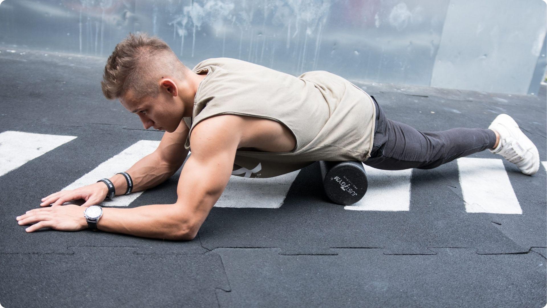 A man in a sleeveless shirt and workout pants uses a foam roller under his left hip flexor while holding a forearm plank position outdoors.