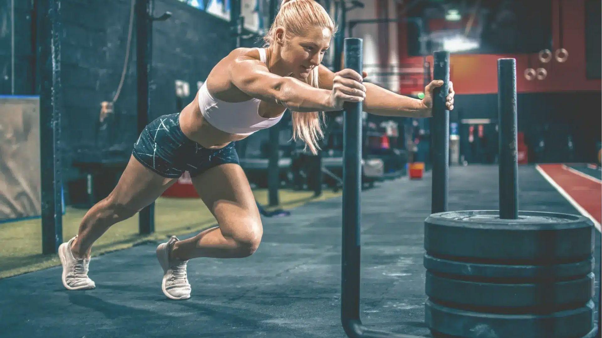 Woman in white sports bra pushing weighted sled in gym with arms extended, driving legs for power sled push workout.