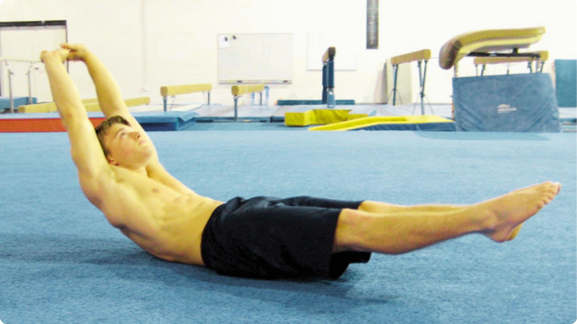 A man in black shorts holds a classic gymnastics hollow body position on a blue mat in a gym.