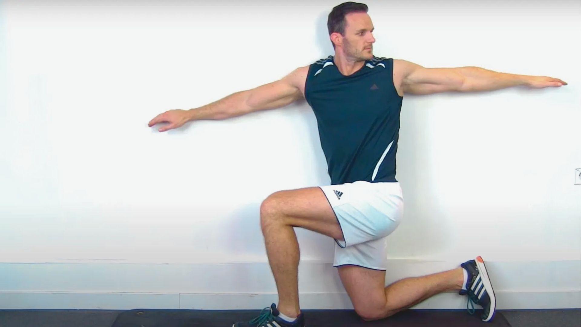 Man in black tank and white shorts kneels by a wall with arms extended in a T-shape.
