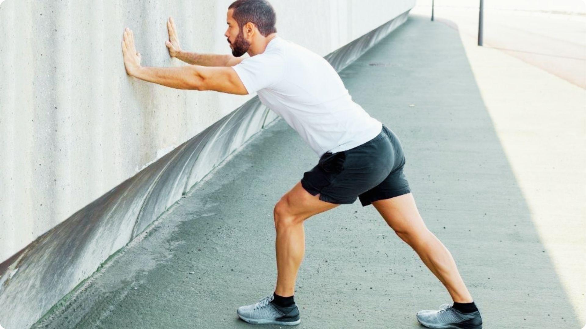 A man in a white t-shirt and black shorts performs a calf stretch against a concrete wall outdoors.
