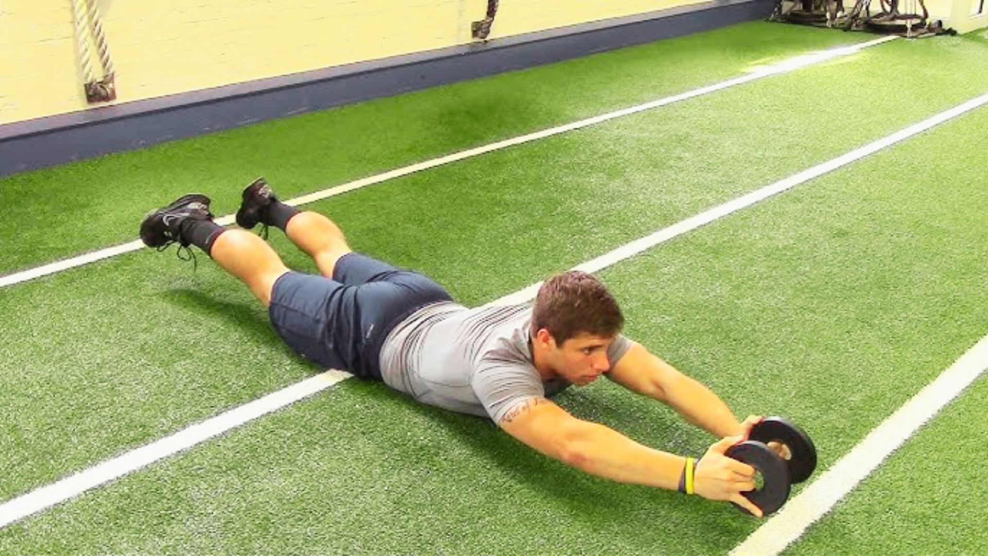 Man lying prone on turf holding weight plates with arms extended overhead.