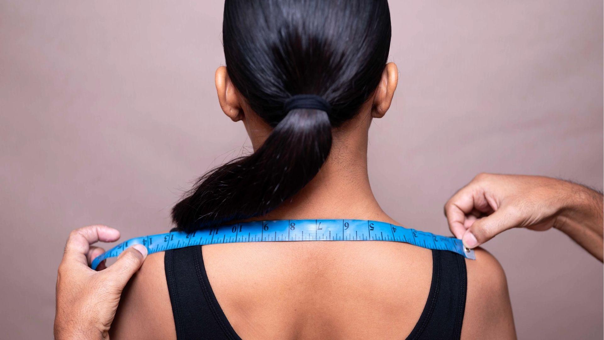 Back view of a woman with hair in a ponytail as hands hold a blue measuring tape across her upper back.