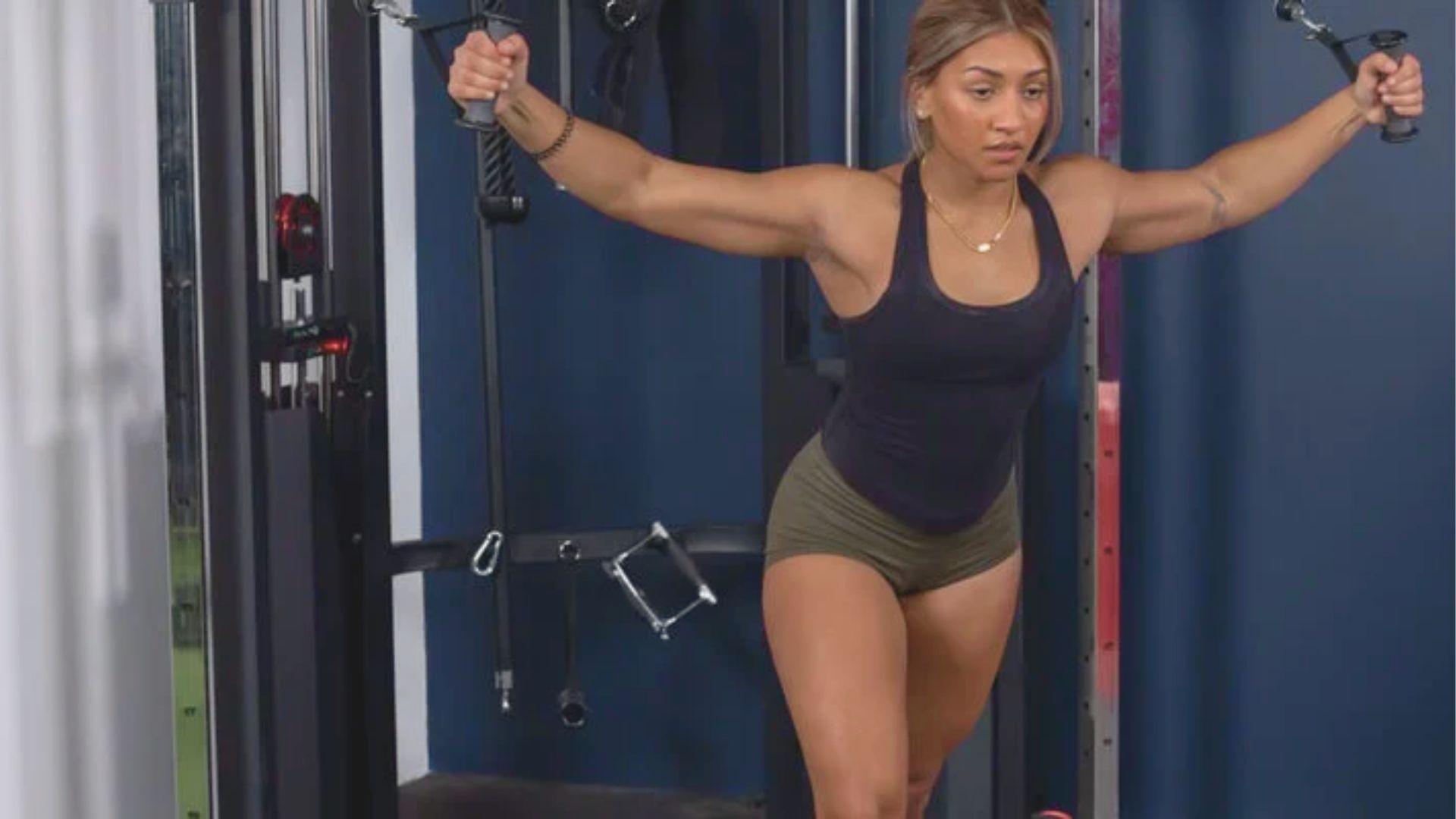 Woman in a navy tank top and olive shorts performing a standing cable fly with arms wide open.