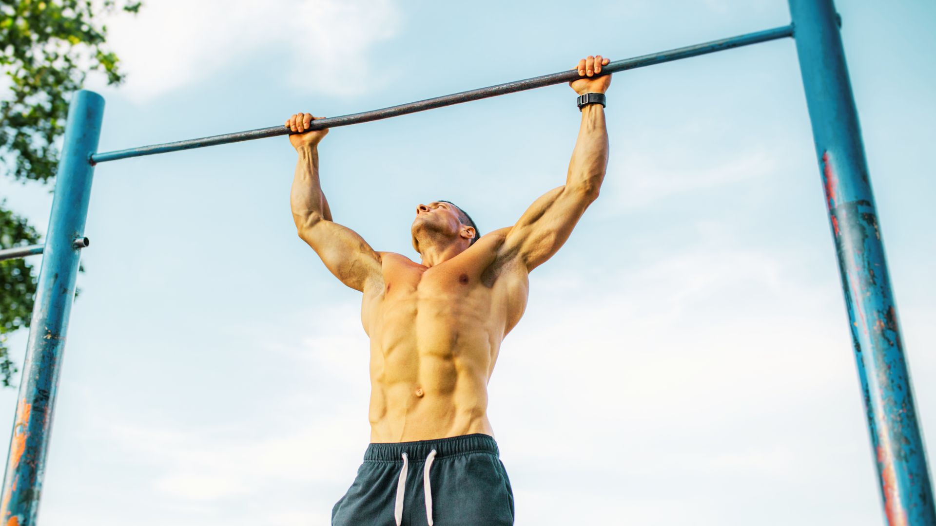 A muscular man performs a wide-grip pull-up on an outdoor blue pull-up bar under a clear sky.
