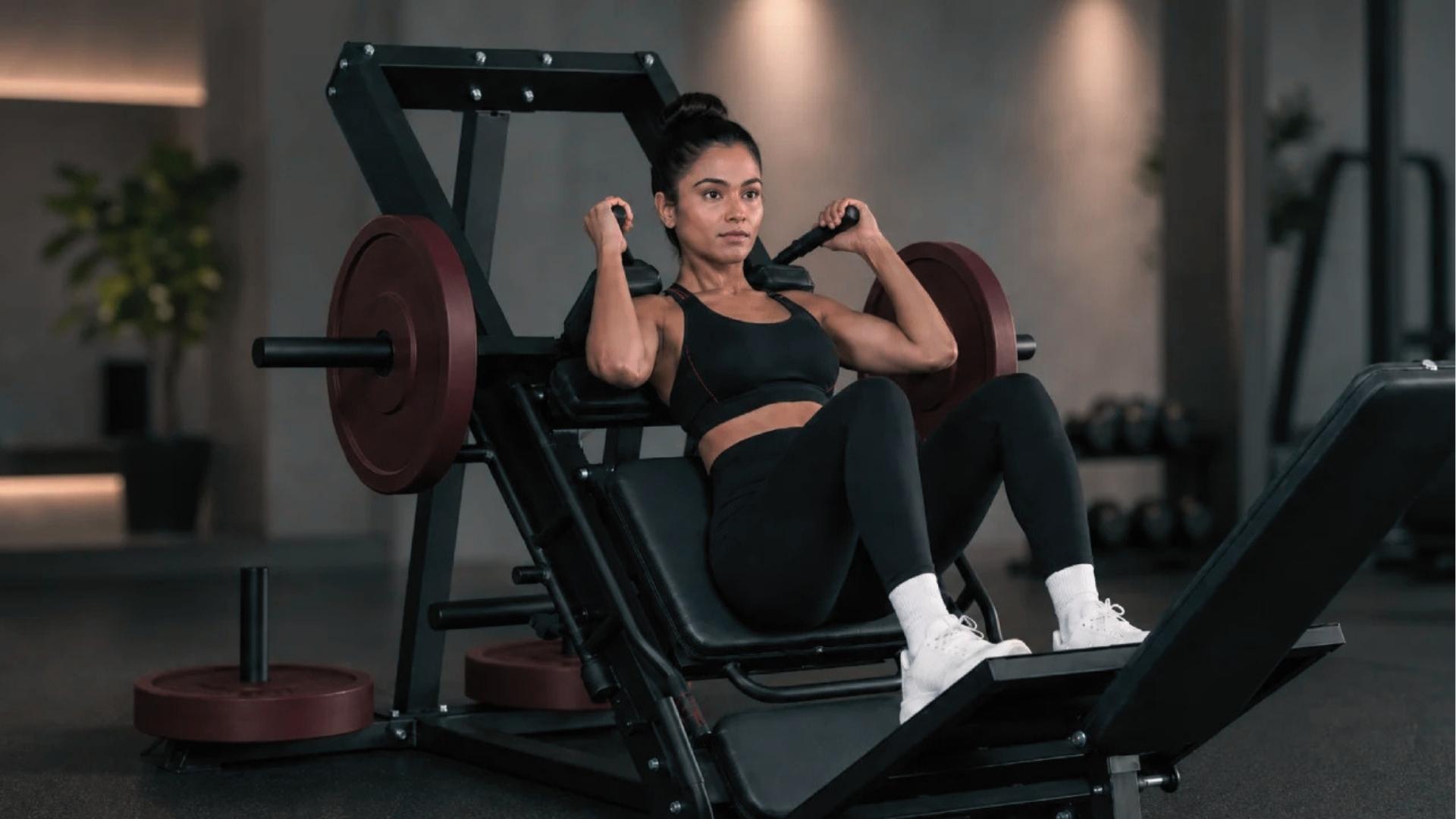 Fit woman in black sports bra and leggings does hack squat with feet low on platform, targeting quadriceps.