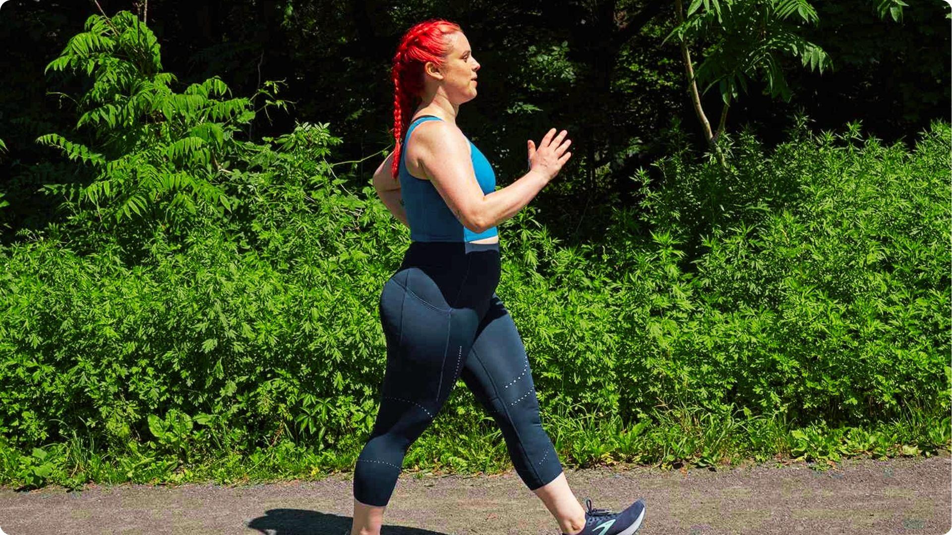 A woman with red braided hair power walks on a paved path lined with green bushes.