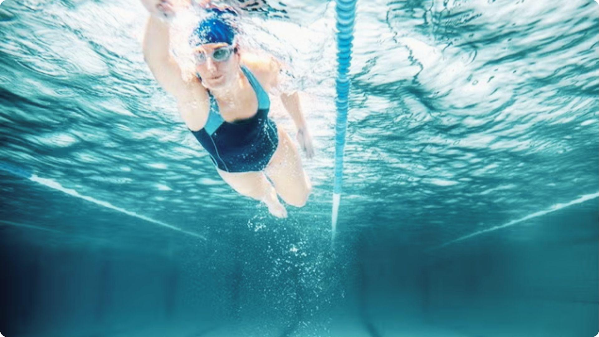 A swimmer in a blue cap and goggles does a freestyle stroke underwater in a pool lane.