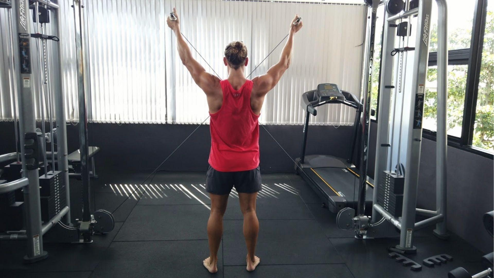 Man in a red tank top performs cable Y raise with arms extended overhead at the cable machine.