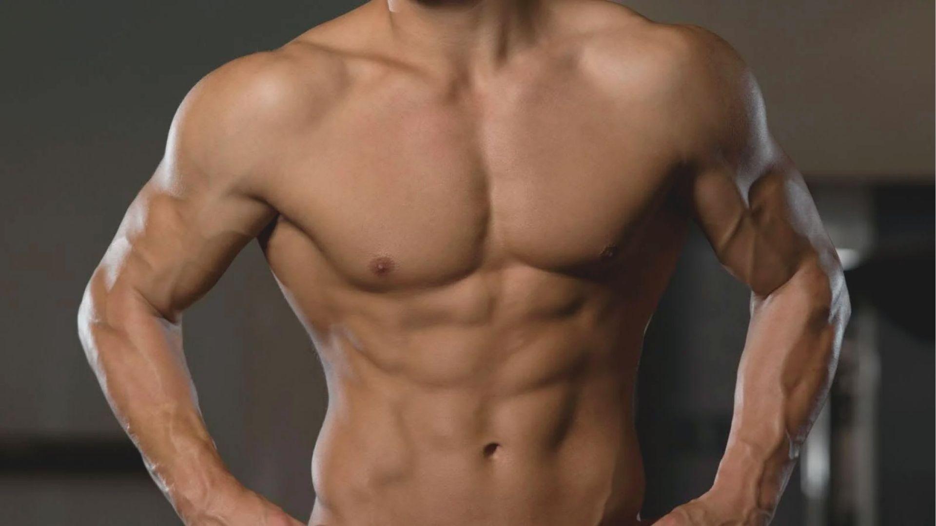 Shirtless male torso showing well-defined chest, shoulders, and abs in a gym setting.