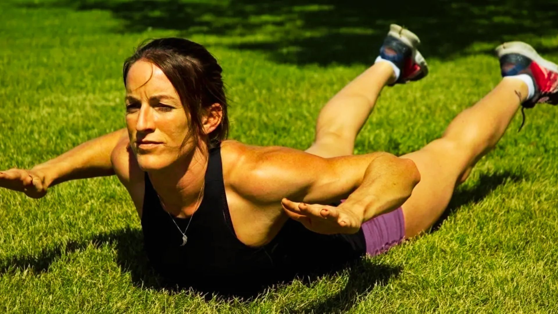 A woman in a black tank top and purple shorts performs a superman hold on grass, lifting her chest and legs off the ground.