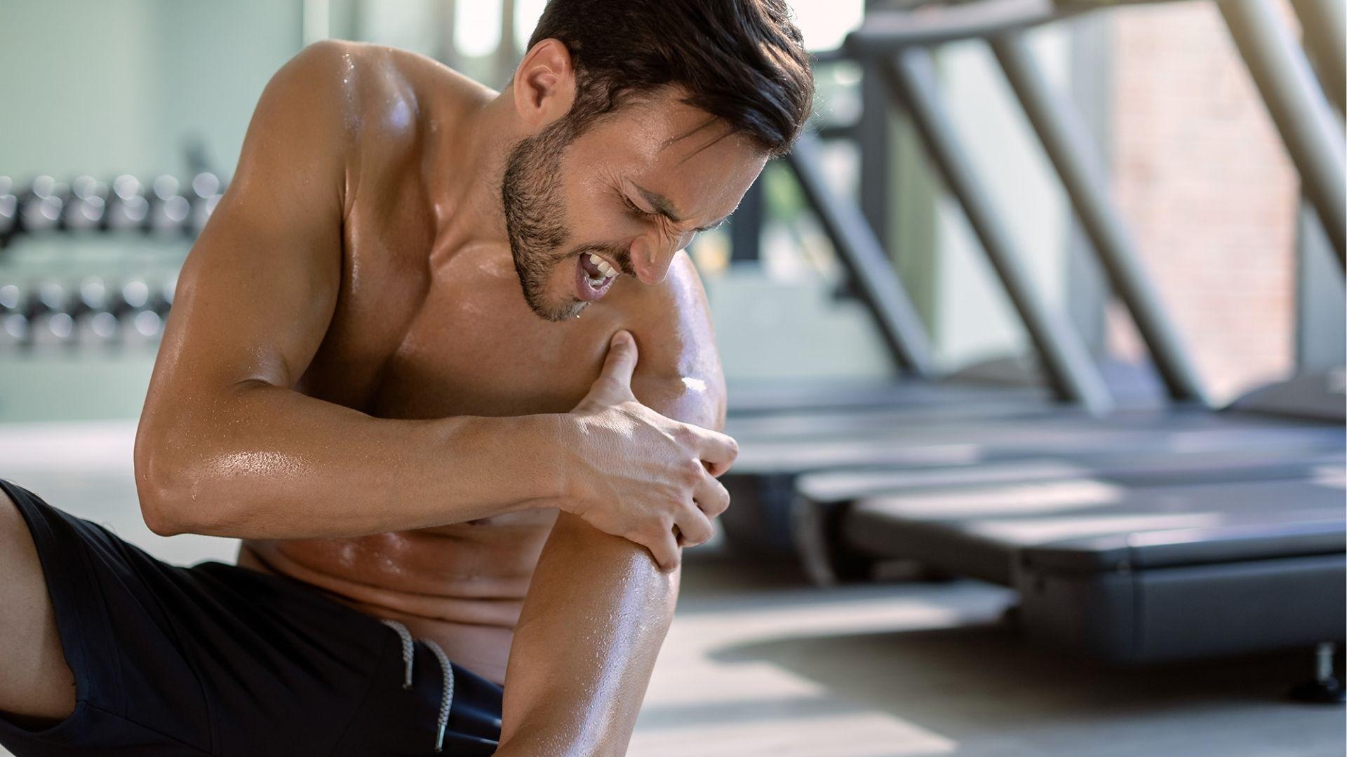 Shirtless man in a gym grimaces while clutching his left shoulder, showing signs of discomfort.