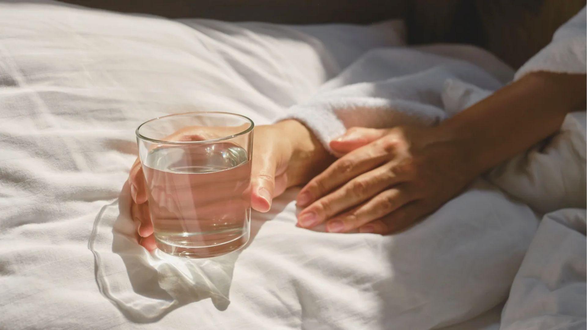 Close-up of hands holding a glass of water while resting on white sheets in sunlight.