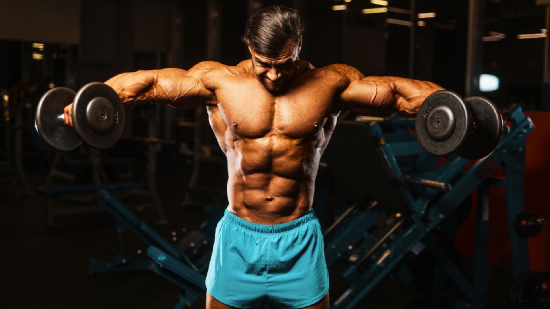 Muscular man in teal shorts does dumbbell lateral raises, showcasing shoulder definition.