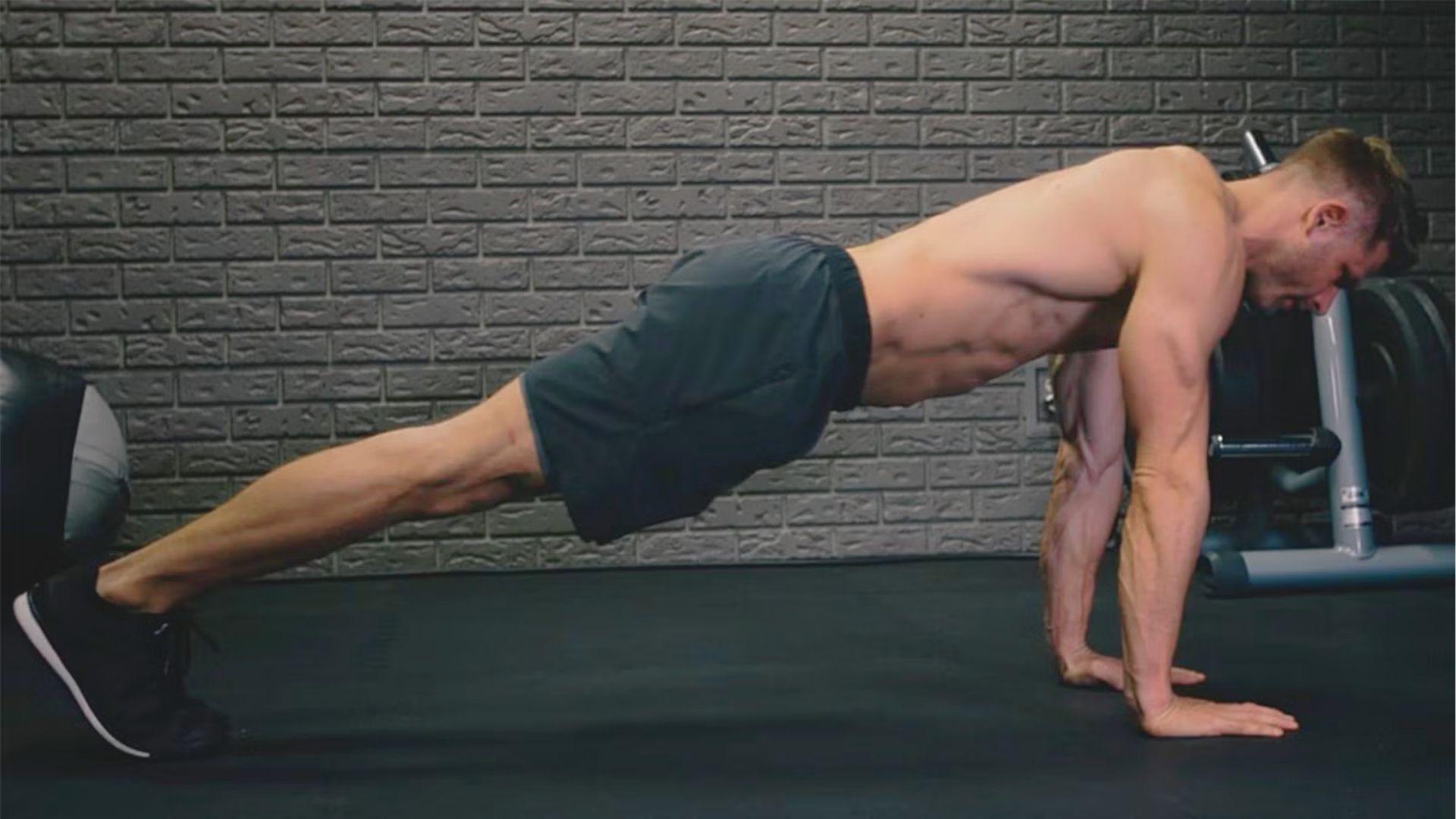 Shirtless man in black shorts holding a plank position with straight arms in a gym with a brick wall background.