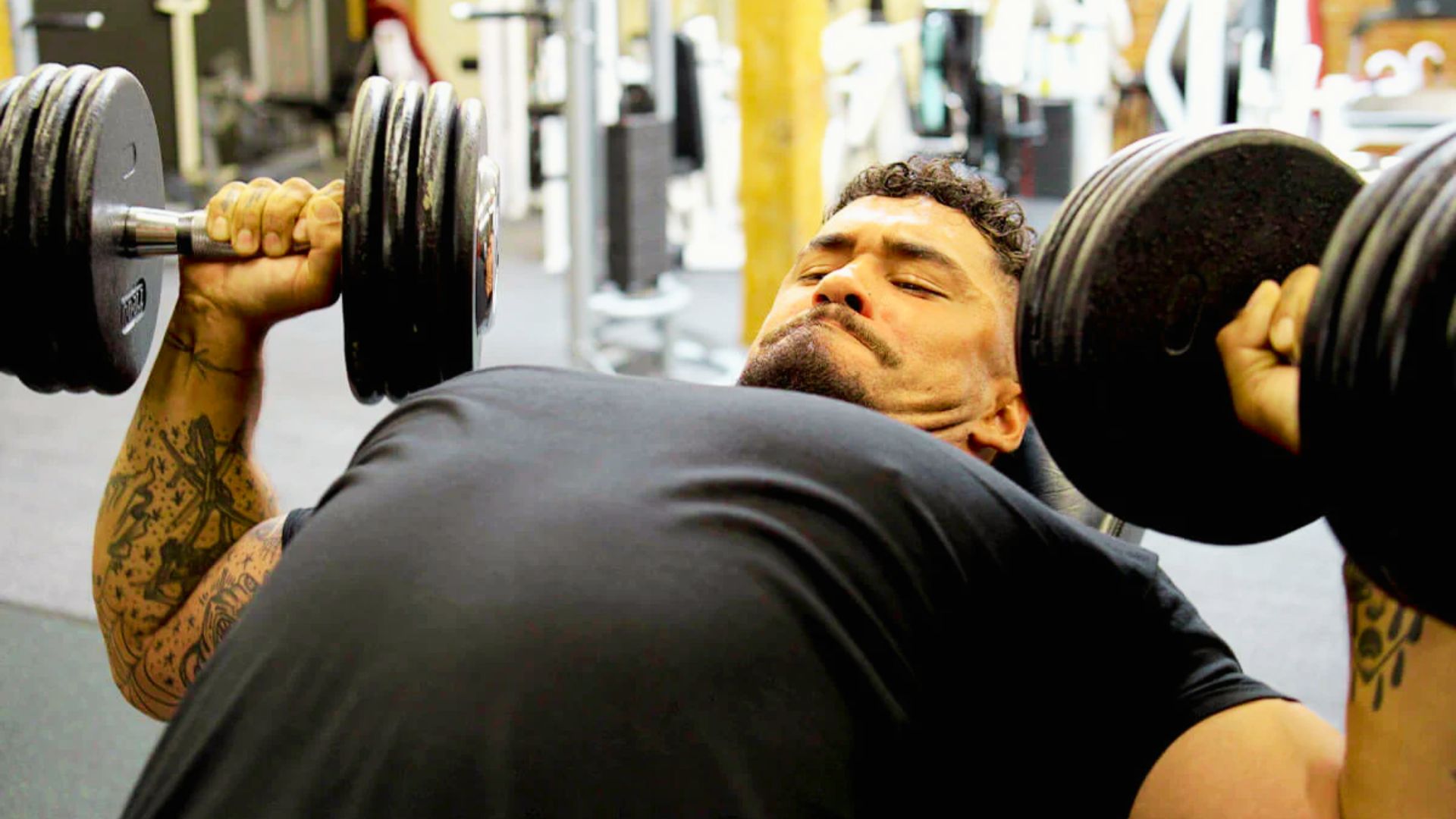 Tattooed man in black shirt presses heavy dumbbells while lying on a gym bench.
