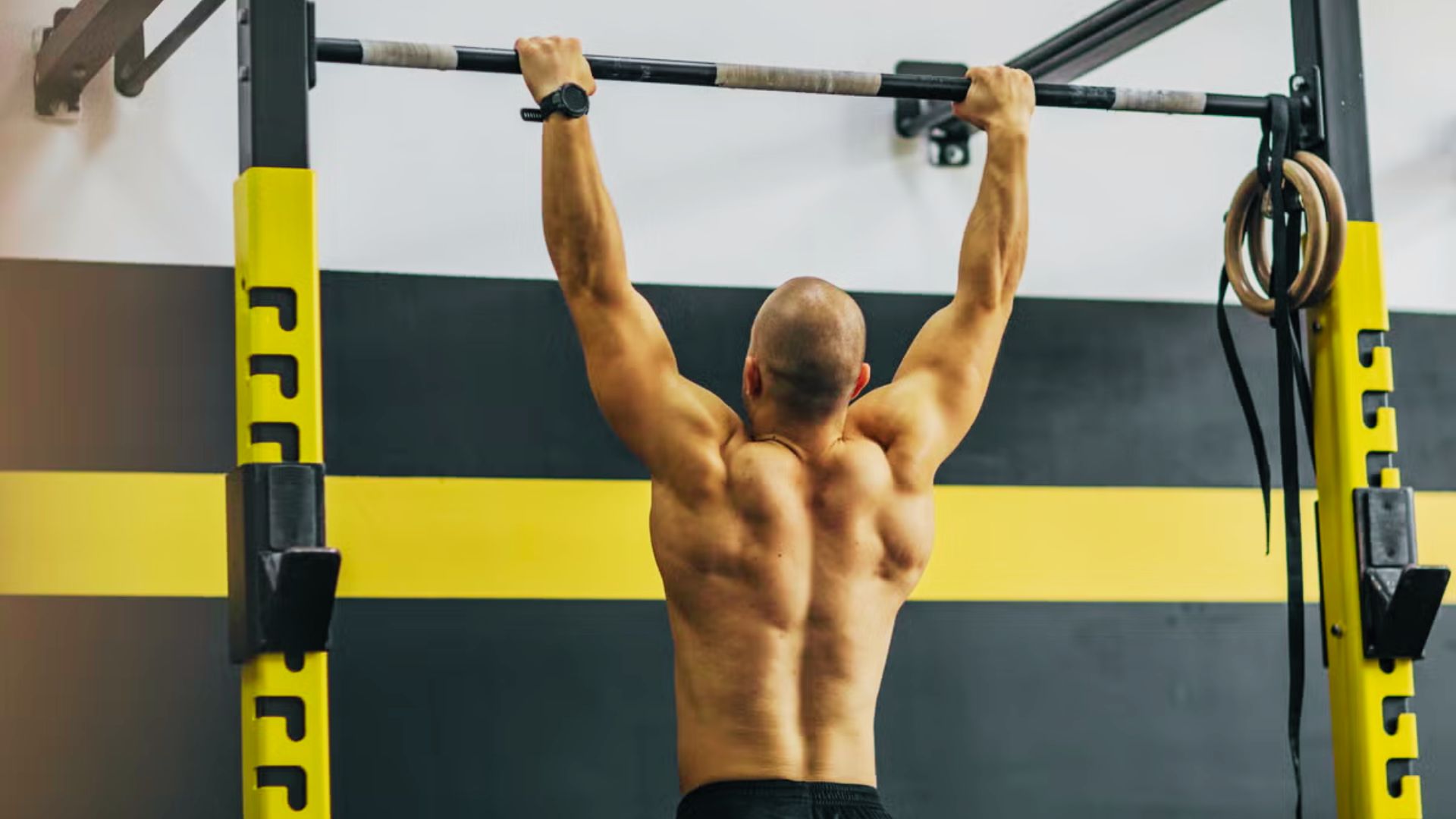 Shirtless man grips a pull-up bar, displaying a strong, defined back in the gym.