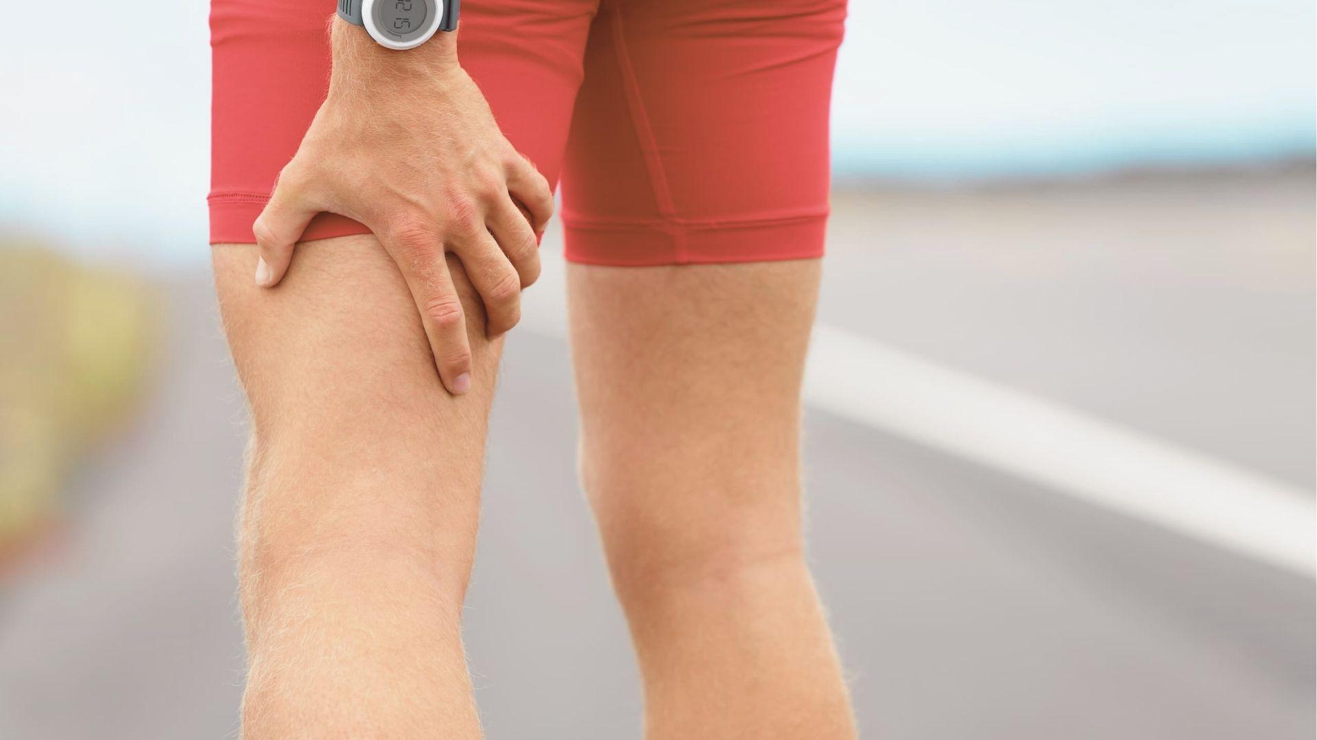 Athlete in red shorts stands on a road, gripping the back of his thigh with a pained expression.
