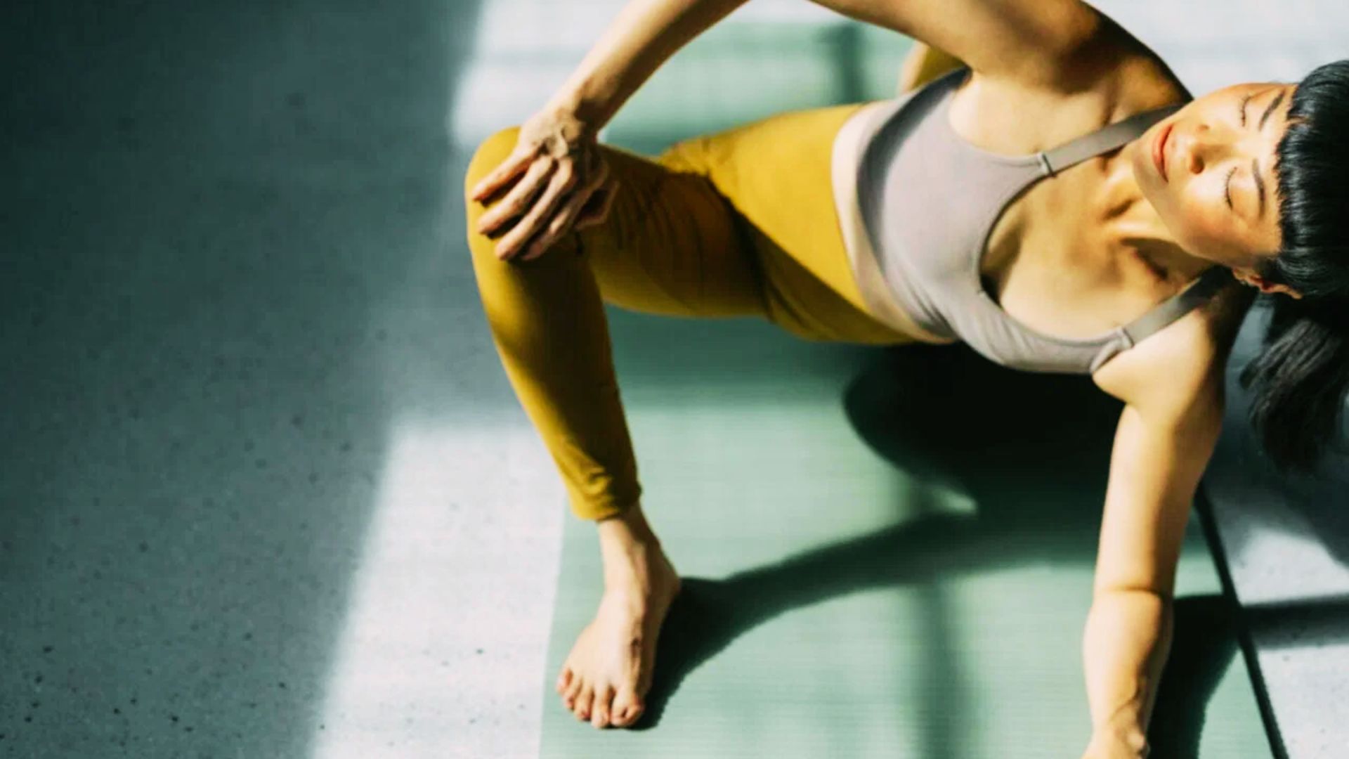 Woman in yellow leggings and grey sports bra performs a side stretch on a yoga mat in sunlight.