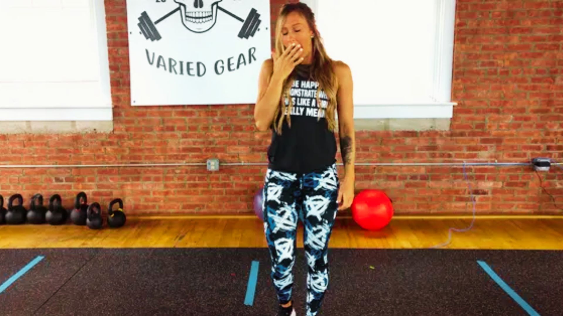 A woman in a gym stands in front of a brick wall with “Varied Gear” signage, yawning with her hand over her mouth.