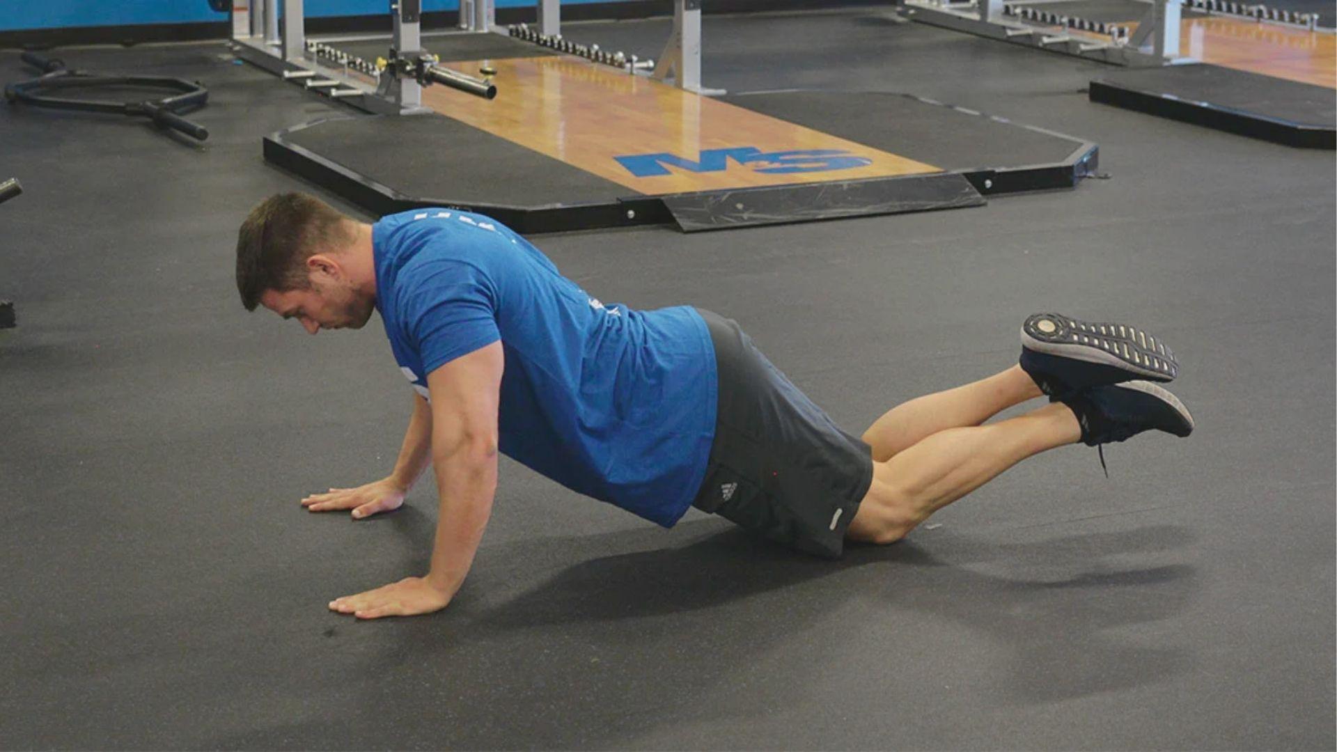 Man in a blue t-shirt performing a knee negative push up on gym flooring with feet crossed.