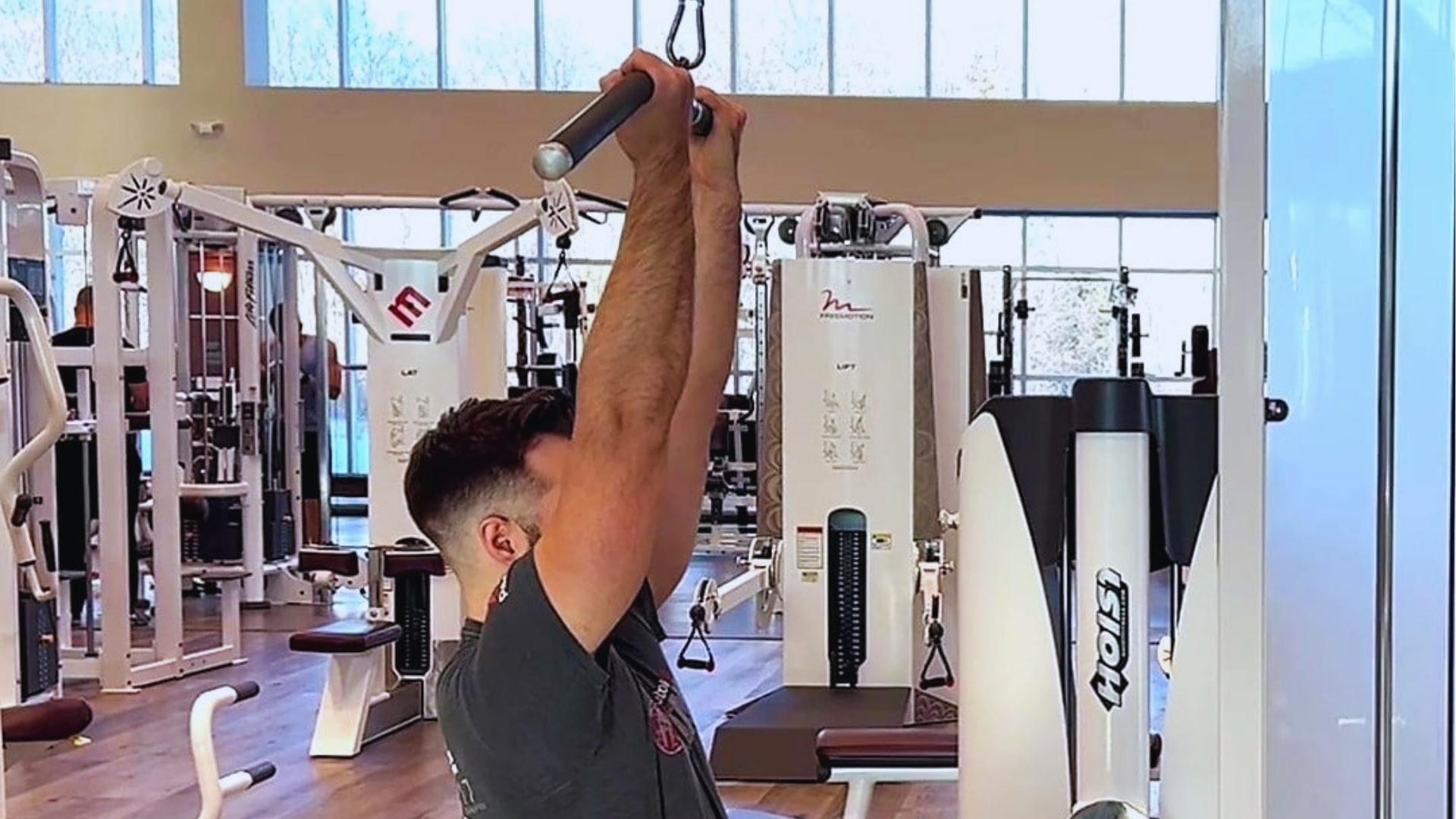 Man in gray shirt grips pulldown bar attachment with neutral grip at top of lat pulldown motion in bright modern gym.
